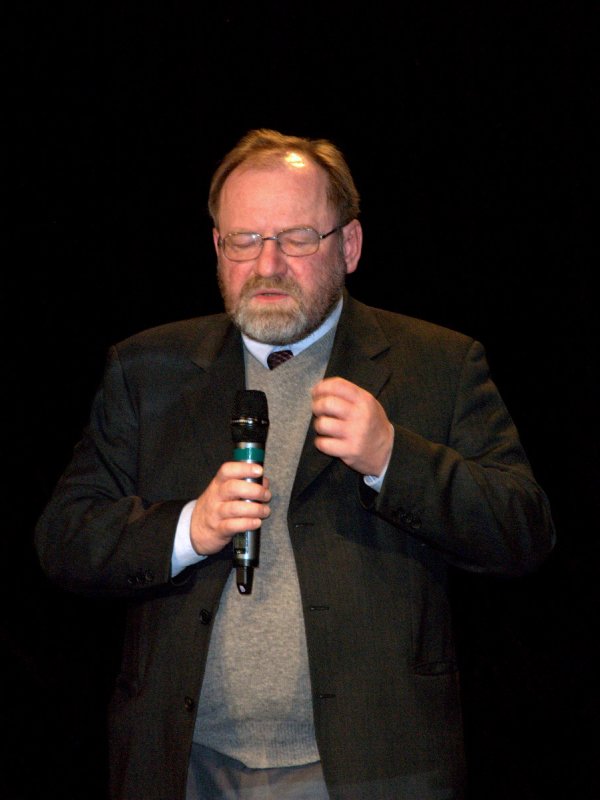 Mężczyzna z brodą i wąsami, w okularach w ciemnej marynarce i szarym swetrze pod spodem, stoi z mikrofonem w ręku.