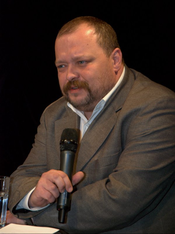 Mężczyzna z brodą i wąsami, w brązowej marynarce, z mikrofonem w ręku.