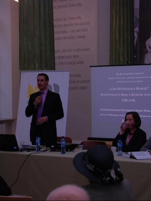 Stół konferencyjny; Richard Butterwick stoi z mikrofonem w ręku; siedzą kobieta i mężczyzna, z tyłu plakaty i ekran.