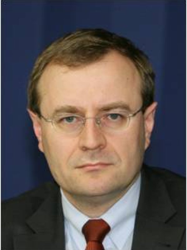 Mężczyzna z jasnymi włosami, w delikatnych okularach, ciemnej marynarce, białej koszuli i wiśniowym krawacie