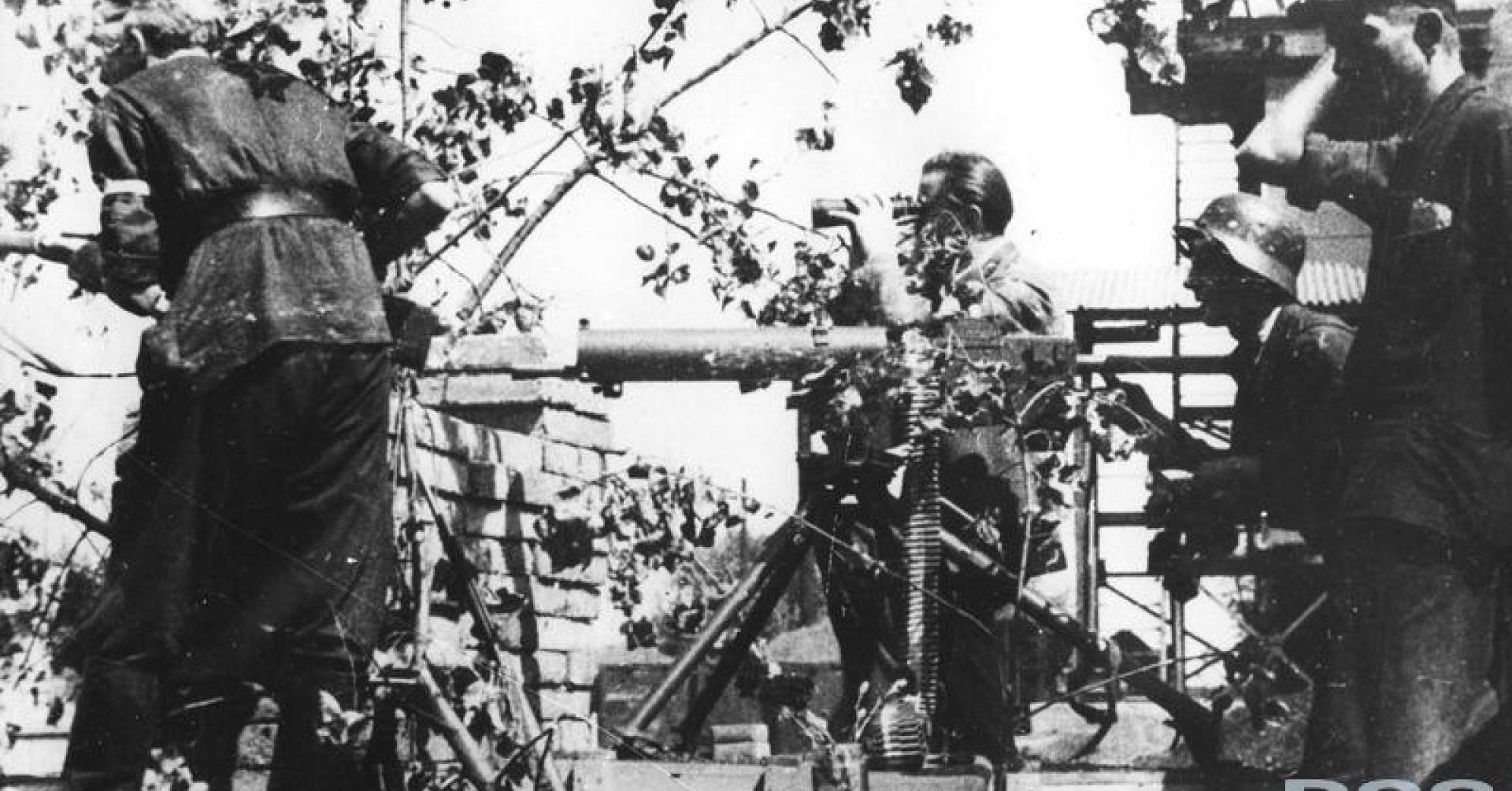 Kobiety, dzieci i mężczyźni z rękami podniesionymi do góry wychodzą z bramy pod nadzorem uzbrojonych żołnierzy niemieckich. Na pierwszym planie kobieta z torbą, odwracająca się do tyłu i chłopczyk w czapce z daszkiem.