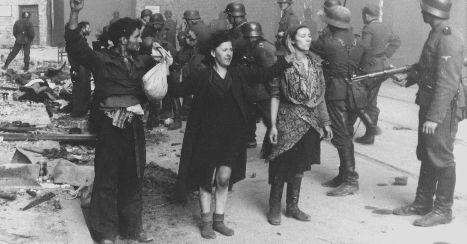 Mężczyzna i dwie kobiety pod nadzorem żołnierza niemieckiego, w tle maszerujący żołnierze