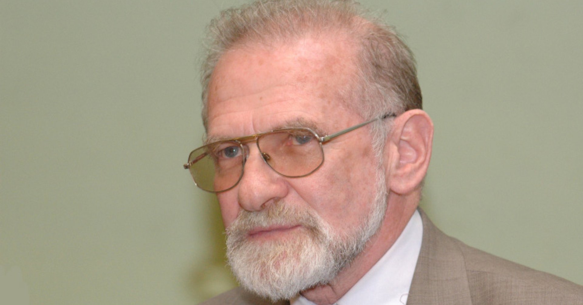 Siwy mężczyzna z brodą i w okularach w beżowej marynarce i ciemnym krawacie.
