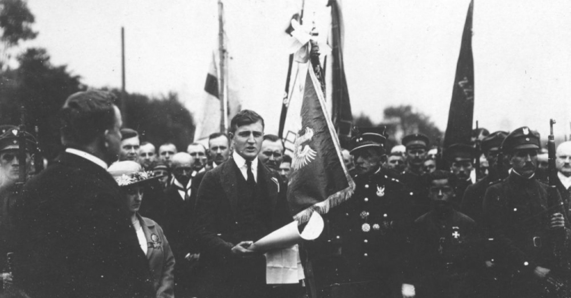 Grupa wojskowych i cywilów trzymających sztandary. Po środku mężczyzna w garniturze, trzymający w ręku arkusz papieru, obok kobieta w kapeluszu