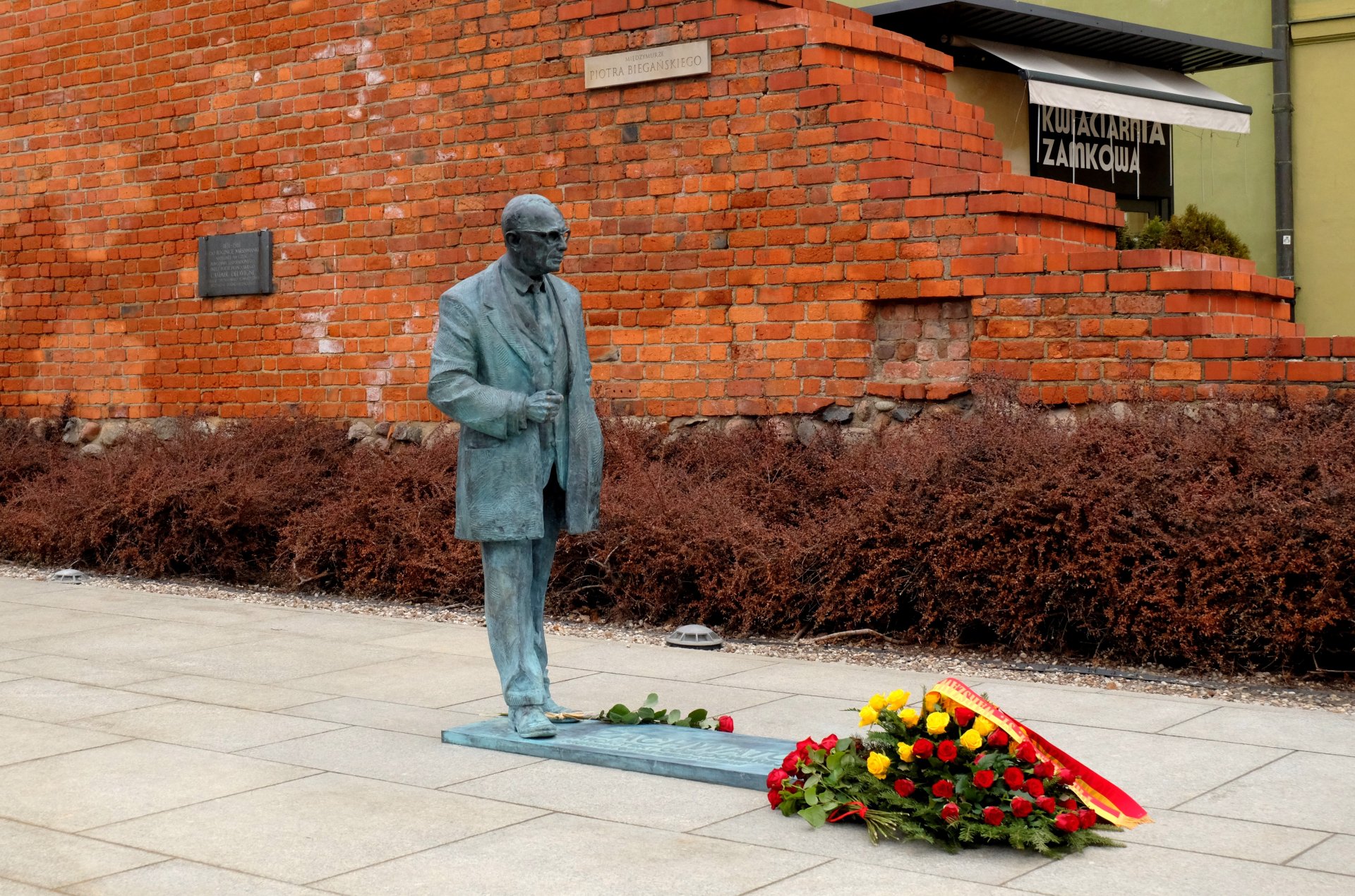 Pomnik przedstawiający stojącą sylwetkę mężczyzny w okularach i płaszczu. U stóp pomnika wieniec. W tle - mur ceglany.