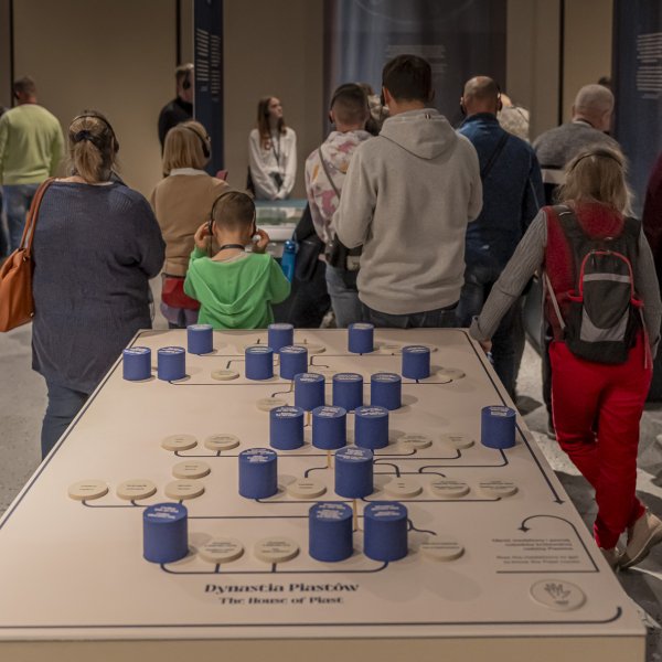 Grupa osób w różnym wieku stoi tyłem do obiektywu. Każdy ma na uszach słuchawki. Na pierwszym planie element wystawy – stół z drzewem genealogicznym dynastii Piastów w formie okrągłych, niebieskich klocków ustawianych w opisanych miejscach.