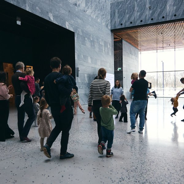 Hol Muzeum. Grupa rodziców z małymi dziećmi  idzie na wystawę czasową. Część osób jest na bosaka. To niedzielne warsztaty dla maluchów.