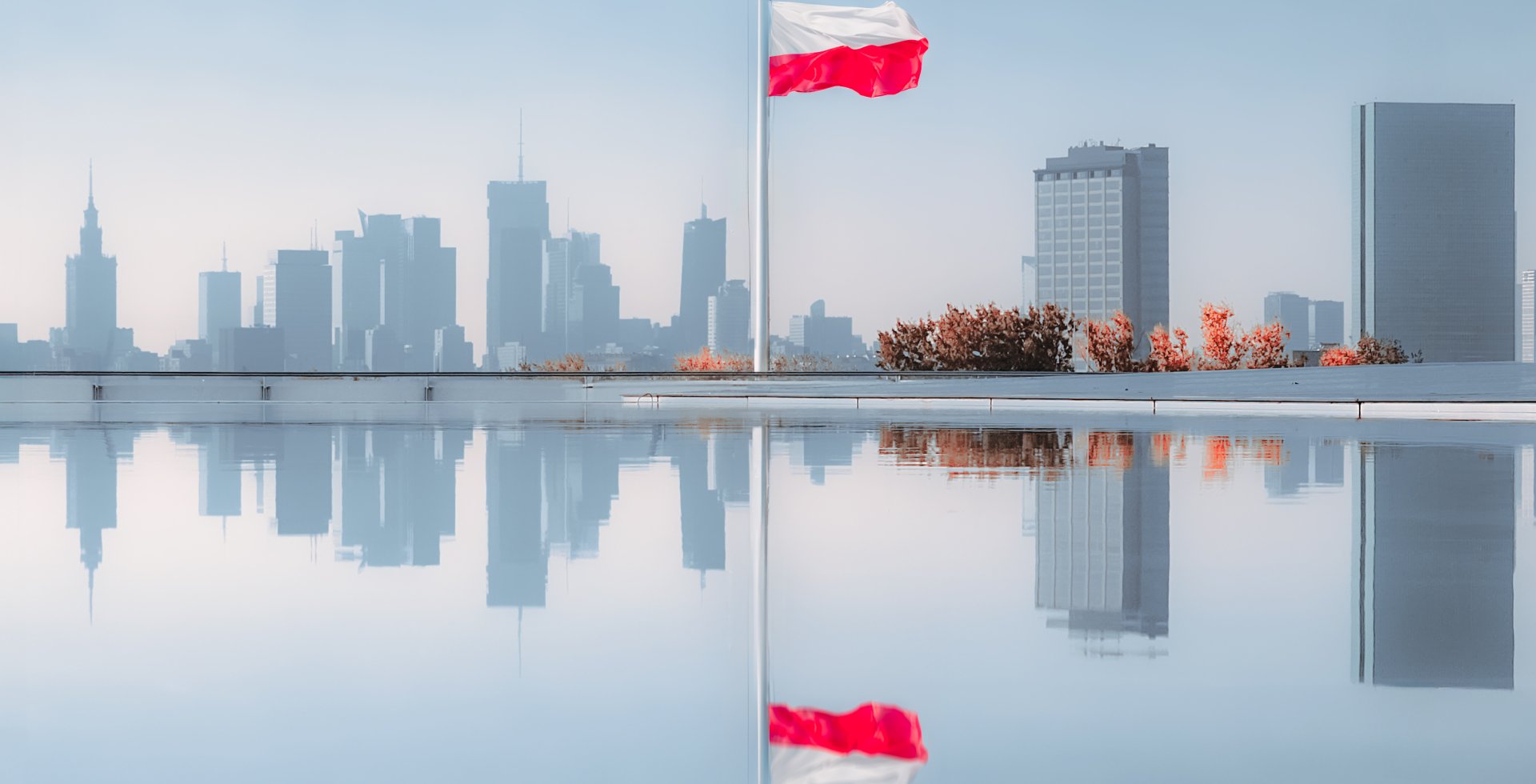 Widok z tarasu na panoramę Warszawy i odbitą flagę w tafli wody.