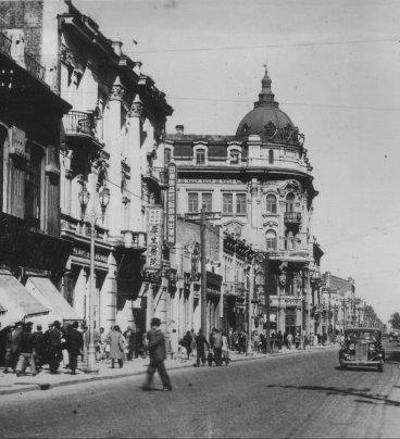 Wielkomiejska ulica z kamienicami, dużo przechodniów na chodnikach. Jezdnią jedzie automobil z początku XX wieku.