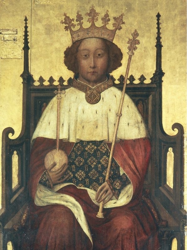 Młody król z jasnymi, kręconymi włosami, w złotej koronie, w płaszczu królewskim, z berłem w lewej i z jabłkiem w prawej dłoni siedzi na drewnianym tronie.