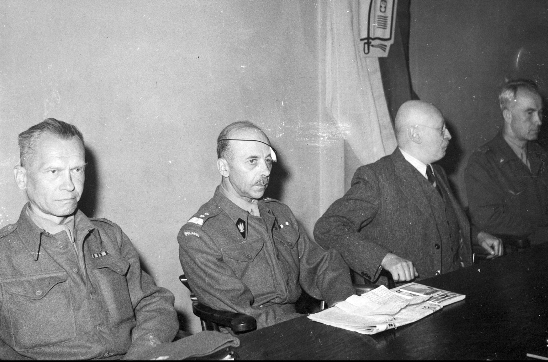 Czterech mężczyzn przy stole, drugi od lewej z opatrunkiem nad lewym okiem. Na ścianie wiszą fotografia i kotwica Polski Walczącej.