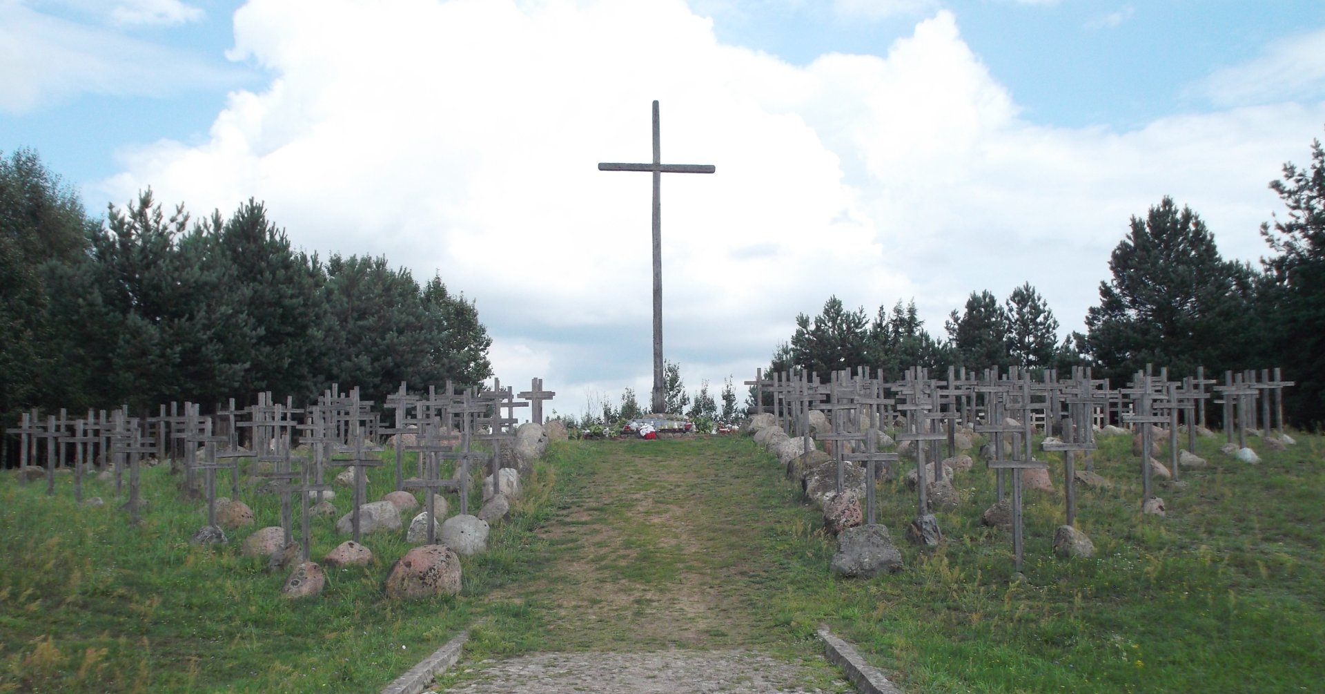 Zdjęcie przedstawia wysoki drewniany krzyż, po obu stronach tego krzyża stoją rzędy mniejszych drewnianych krzyży, a przy nich głazy.