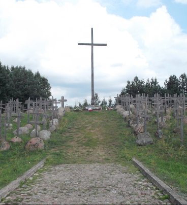 Zdjęcie przedstawia wysoki drewniany krzyż, po obu stronach tego krzyża stoją rzędy mniejszych drewnianych krzyży, a przy nich głazy.