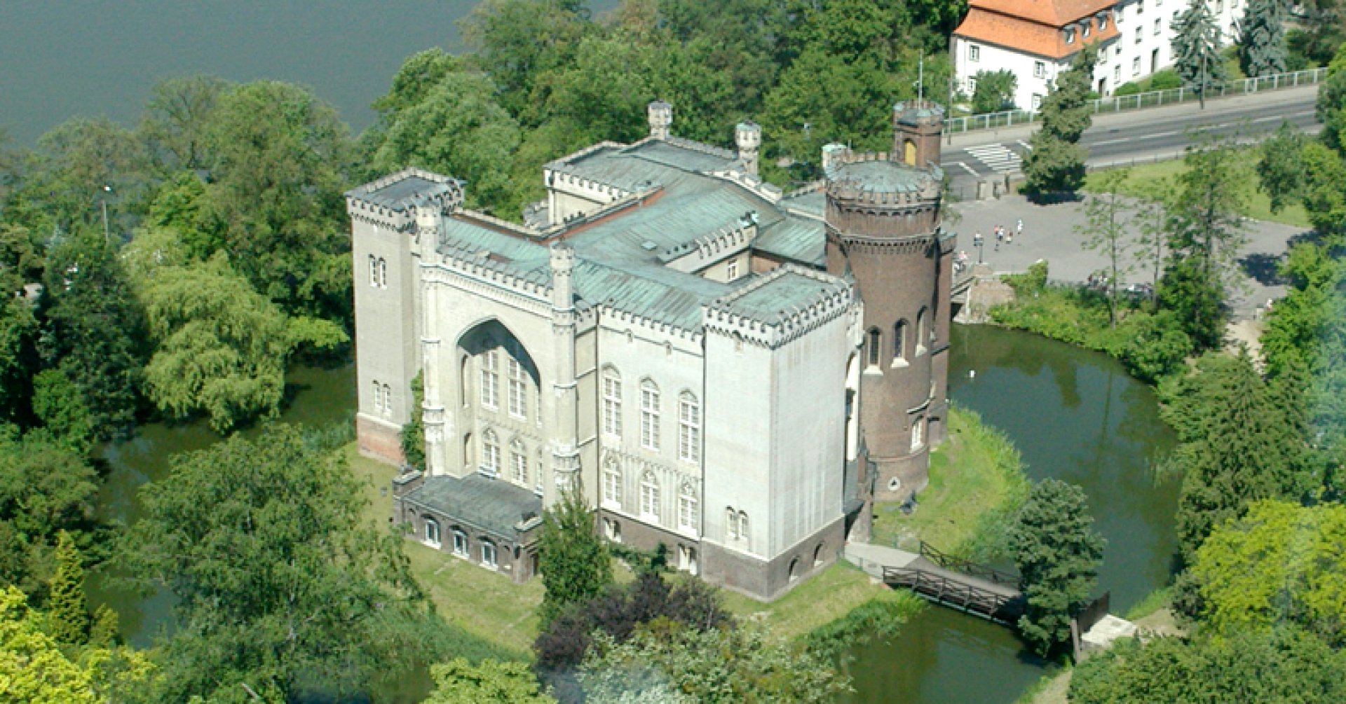 Zdjęcie przedstawia zamek w stylu neogotyckim, otoczony fosą i zielenią drzew.