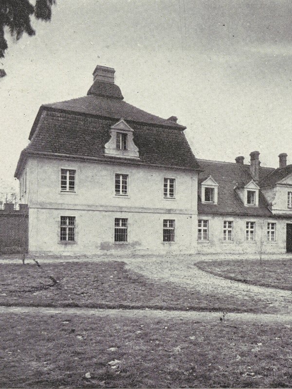 Zdjęcie przedstawia jasny dwukondygnacyjny dworek z wysokim dachem.