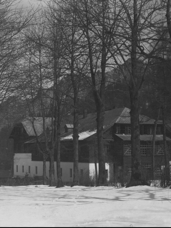 Zdjęcie przedstawia dość duży bydynek w stylu zakopiańskim, otoczony dzrewami. Za budynkiem widać górskie szczyty.