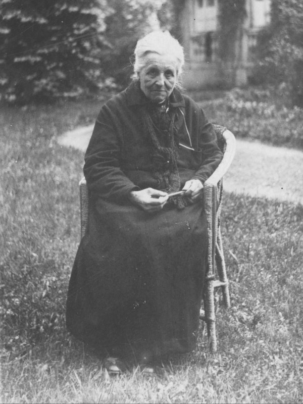 Zdjęcie przedstawia Marię Zamoyską. Staruszka w skromnej czarnej sukni, z siwymi włosami zaczesanymi do tyłu, siedzi na krześle w ogrodzie. 