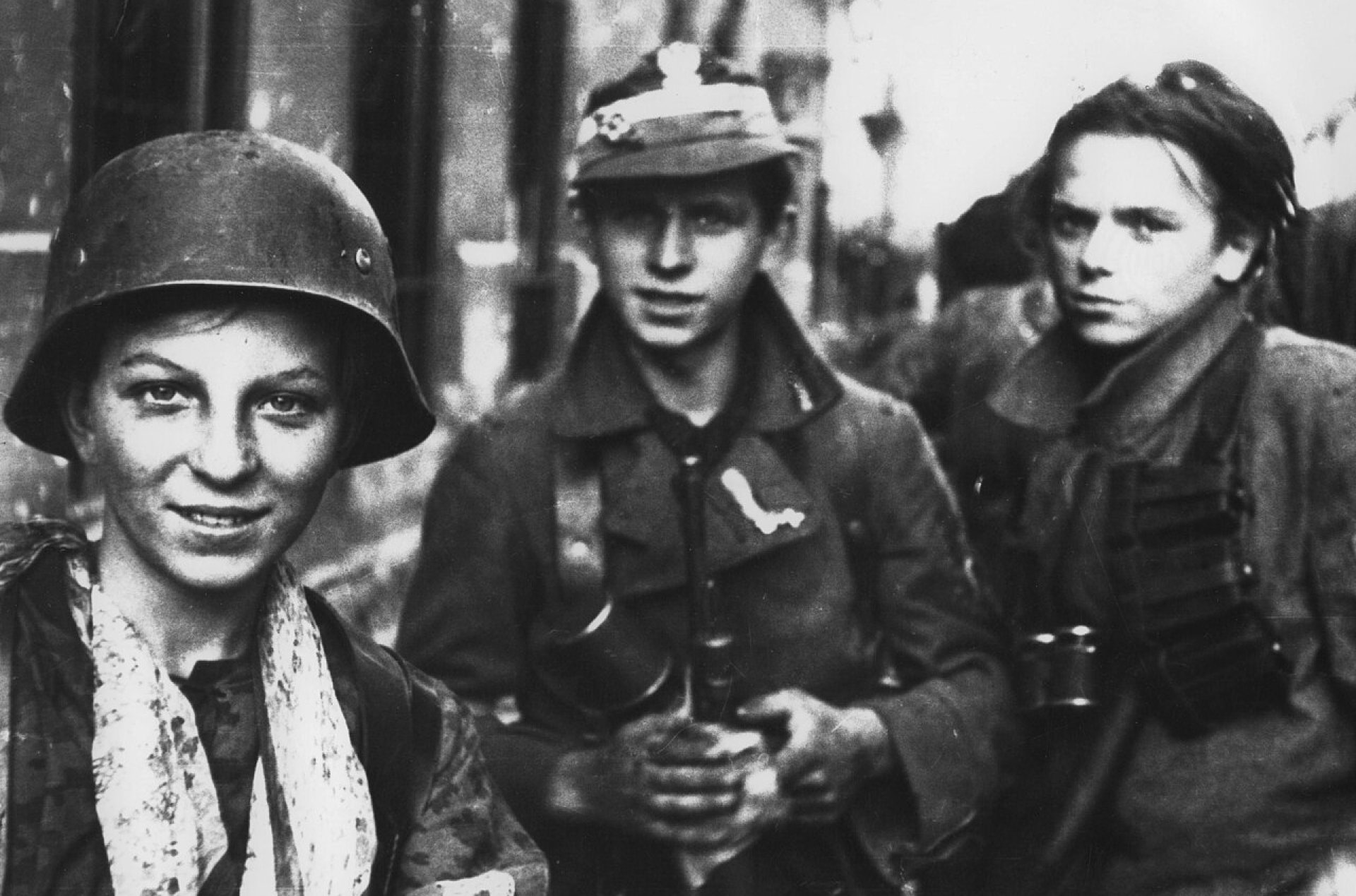Trzech chłopców w umundurowaniu powstańczym. W tle zniszczone budynki Warszawy