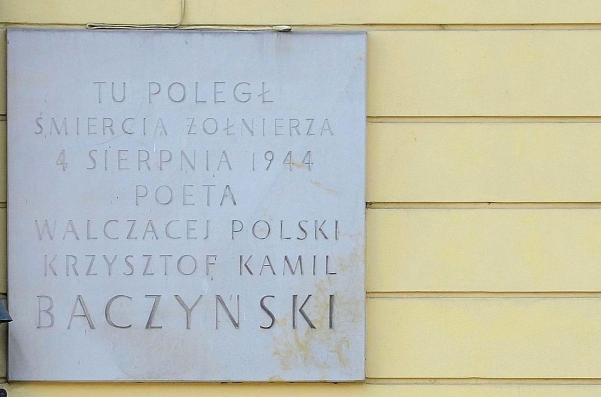 Szara tablica na żółtej ścianie z wygrawerowanymi literami.