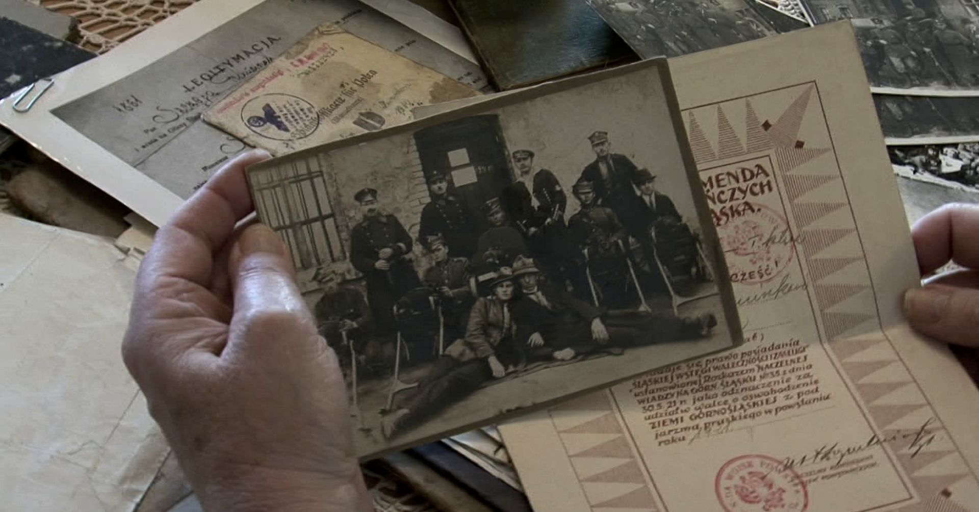 Stary dokument i czarnobiałe zdjęcie w dłoniach. W tle na blacie wiele dokumentów i starych fotografii.