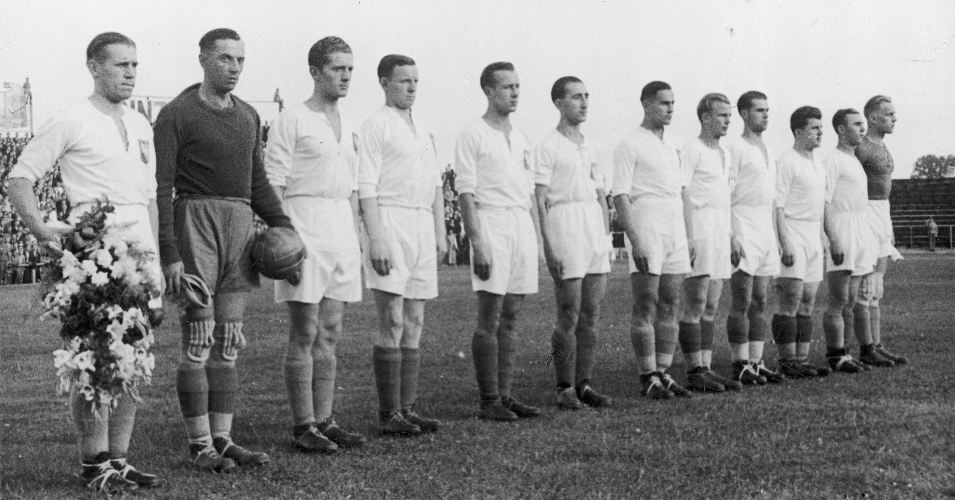 Zdjęcie przedstawia dwunastu piłkarzy stojących w szeregu na stadionie. Pierwszy z lewej trzyma w ręce wieniec kwiatów.