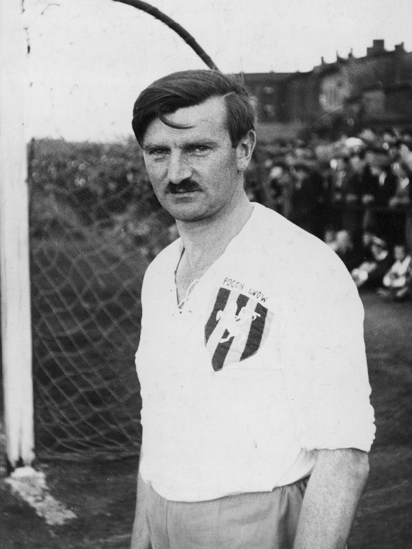 Zdjęcie przedstawia Wacława Kuchara - mężczyznę w średnim wieku, z wąsami, z ciemnymi włosami zaczesanymi na bok, w białej koszulce z logiem klubu Pogoń Lwów.