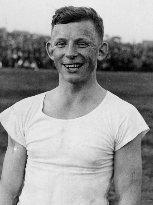 Zdjęcie przedstawia Ernesta Wilimowskiego - młodego, roześmianego mężczyznę o piegowatej twarzy i odstających uszach, w białej, opiętej koszulce. W tle - boisko. 