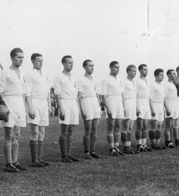 Zdjęcie przedstawia dwunastu piłkarzy stojących w szeregu na stadionie. Pierwszy z lewej trzyma w ręce wieniec kwiatów.