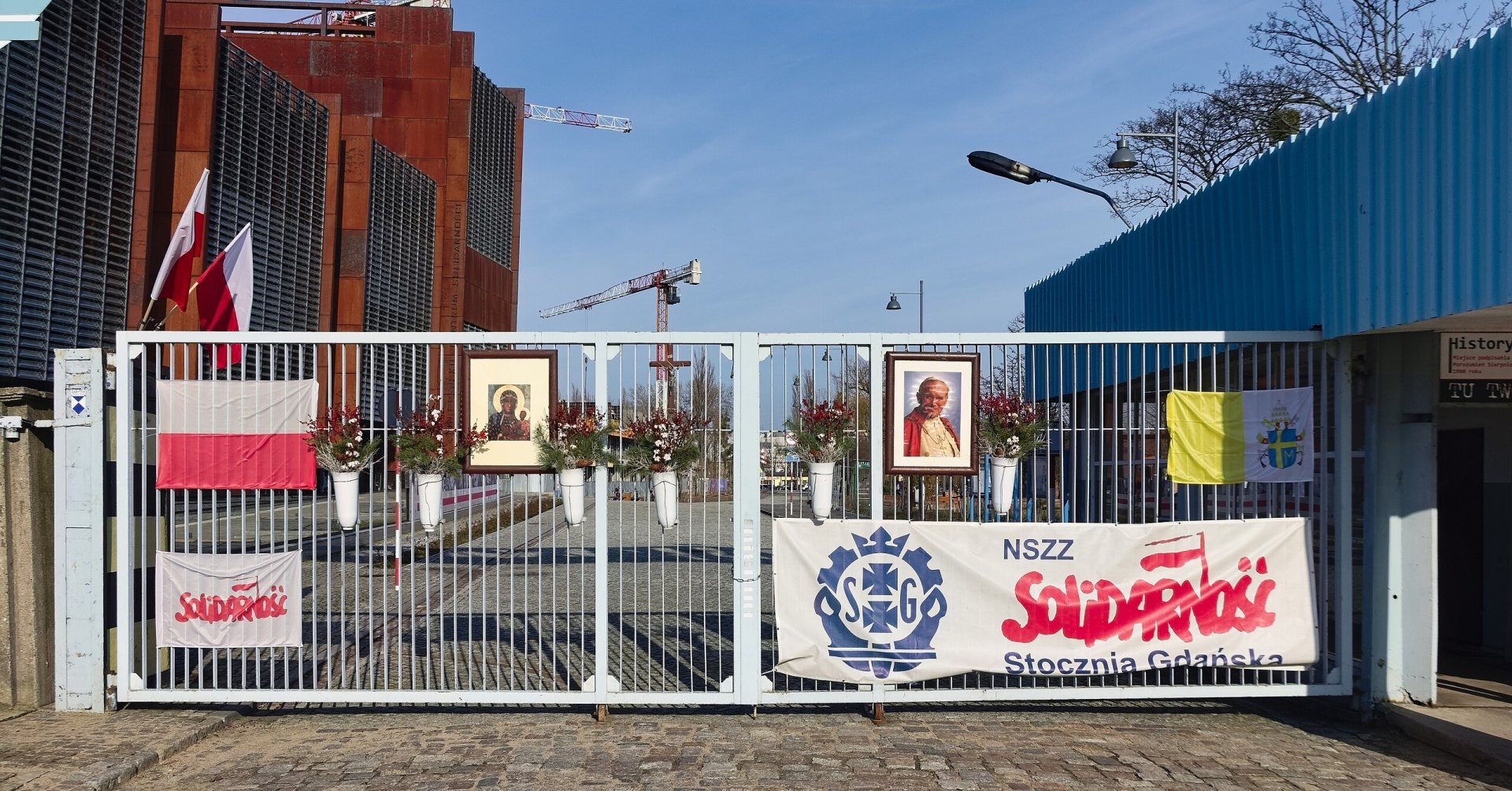 Zdjęcie przedstawia bramę z wiszącymi na niej transparentami „NSZZ Solidarność”, flagami polską i papieską, obrazem Matki Boskiej Częstochowskiej i portretem Jana Pawła II. 