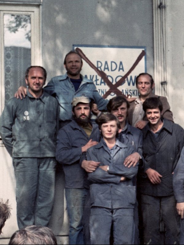 Zdjęcie przedstawia grupę robotników w niebieskich ubraniach roboczych. Robotnicy pozują do zdjęcia na tle tablicy z przekreślonym napisem „Rada Zakładowa”.