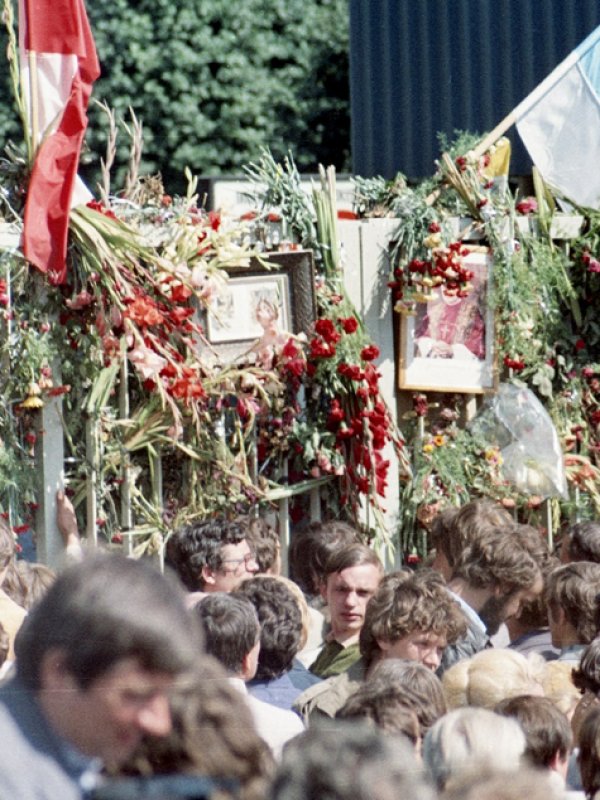 Zdjęcie przedstawia tłum ludzi zgromadzonych przed przystrojoną kwiatami i flagami bramą.