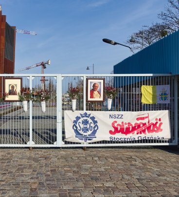 Zdjęcie przedstawia bramę z wiszącymi na niej transparentami „NSZZ Solidarność”, flagami polską i papieską, obrazem Matki Boskiej Częstochowskiej i portretem Jana Pawła II. 