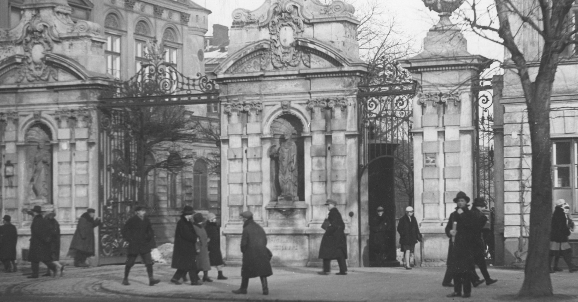Zdjęcie przedstawia zabytkową bramę ozdobioną rzeźbami. Przed bramą, na ulicy widać kilkunastu przechodniów w ciemnych płaszczach.