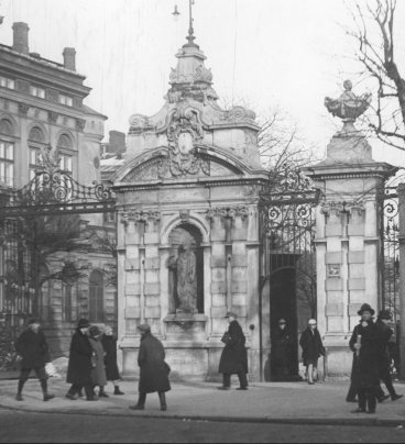 Zdjęcie przedstawia zabytkową bramę ozdobioną rzeźbami. Przed bramą, na ulicy widać kilkunastu przechodniów w ciemnych płaszczach.