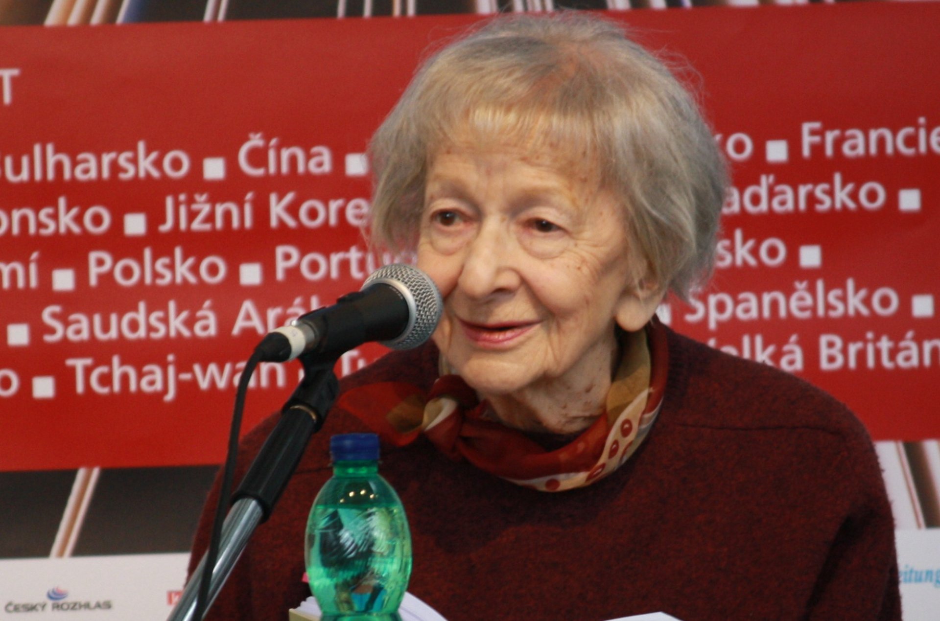 Starsza, siwa kobieta siedzi przed mikrofonem, ma na sobie bordowy sweter, trzyma w rękach rozłożoną książkę. Za nią czerwona i niebieska tablica z białymi napisami.
