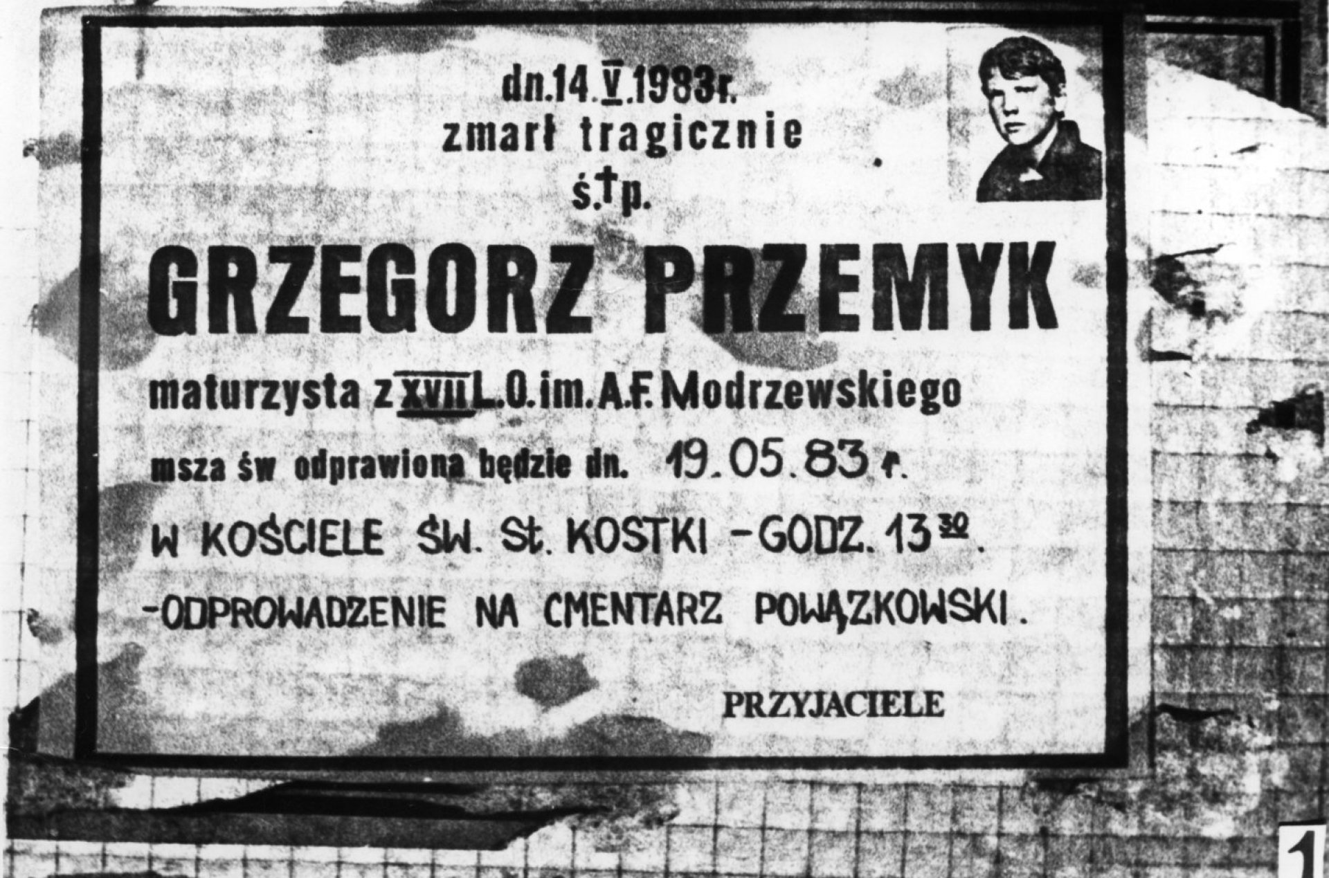 Niekrolog Grzegorza Przemyka ze zdjęciem, powieszony na siatce ogrodzenia. Czarno-biała fotografia