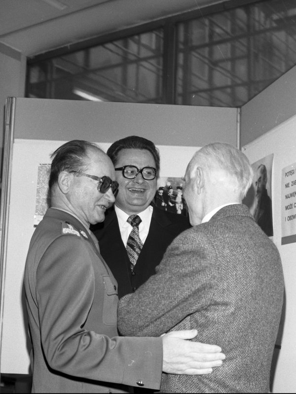 Zdjęcie przedstawia generała Jaruzelskiego w mundurze i ciemnych okularach. Generał rozmawia z dwoma mężczyznami i uśmiecha się do nich.