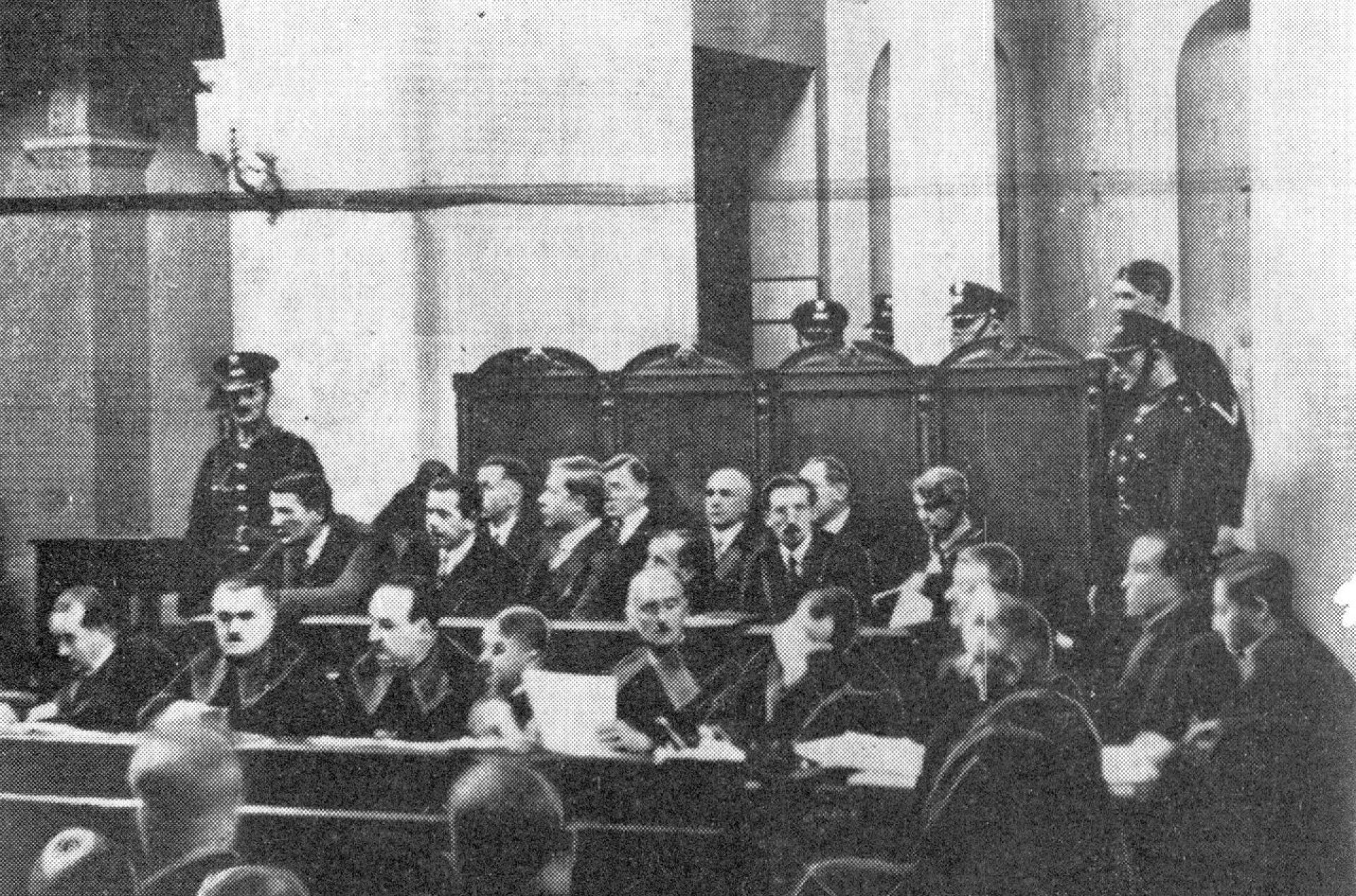 Sala sądowa - widok na mężczyzn siedzących na ławie oskarżonych w otoczeniu policjantów