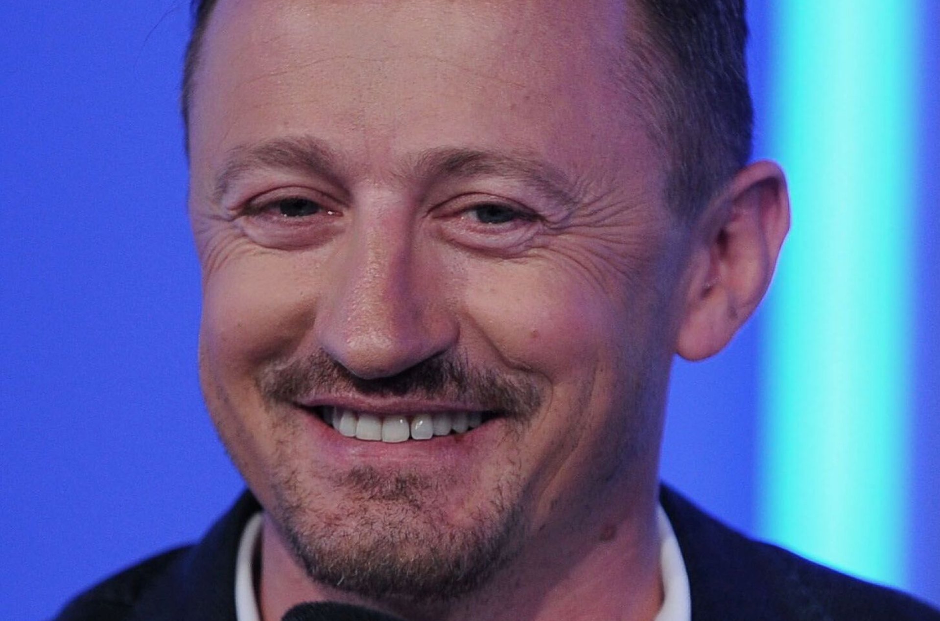 Uśmiechnięty mężczyzna z wąsikiem i bródką siedzi przed mikrofonem.