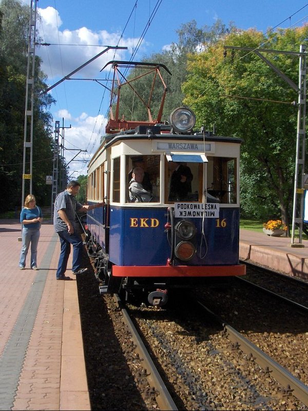 Niebieski wagon WKD kolejki z napisem EKD stojący na peronie małej stacji wśród drzew.