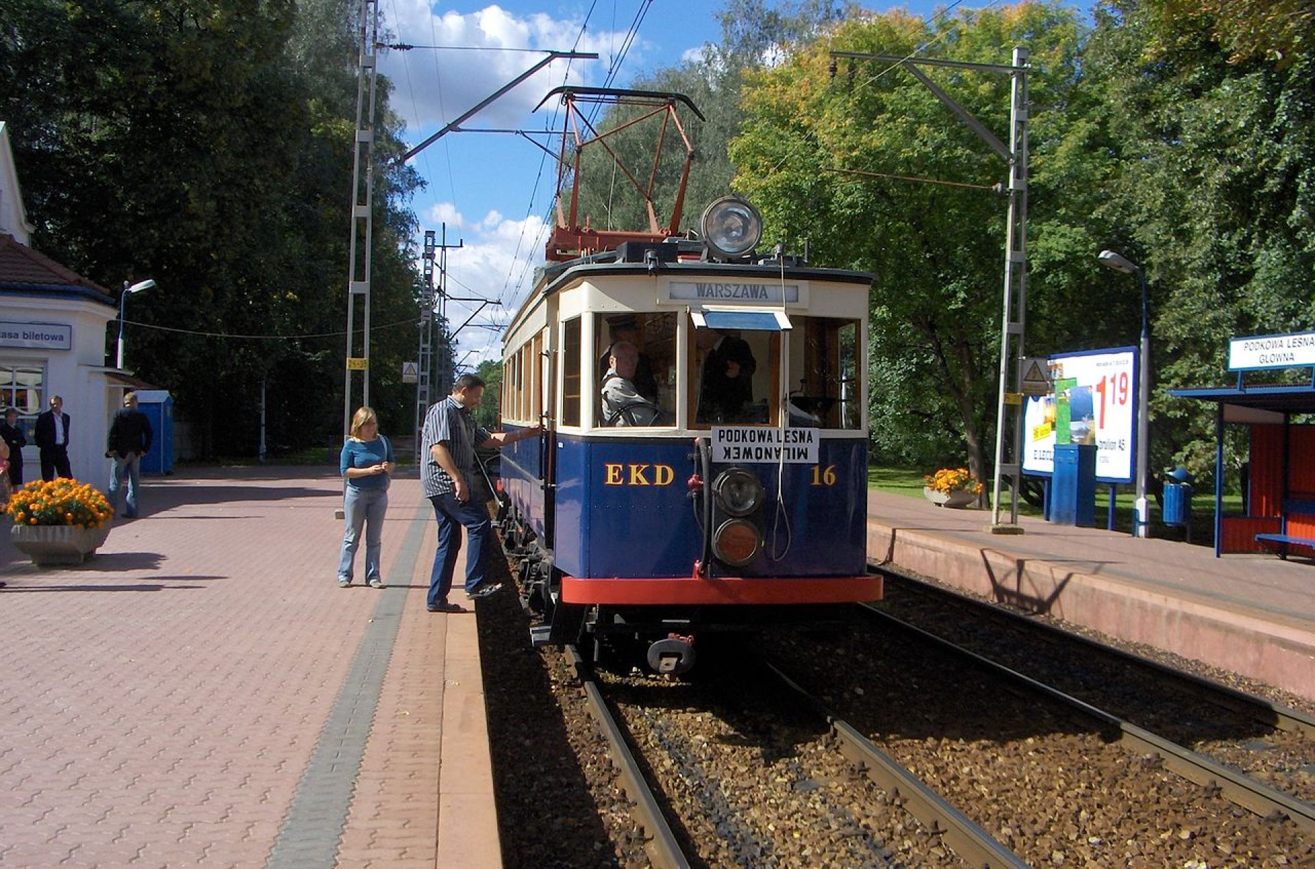 Niebieski wagon WKD kolejki z napisem EKD stojący na peronie małej stacji wśród drzew.