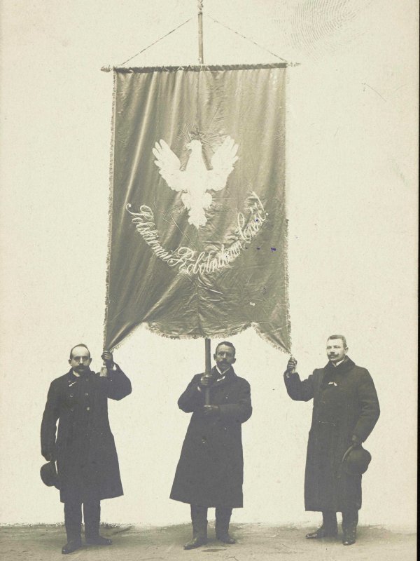 Trzech mężczyzn w płaszczach, z gołymi głowami, trzyma wielki sztandar z Orłem Białym. 