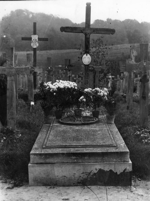 Kamienny nagrobek z drewnianym krzyżem. Na płycie nagrobka donice z chryzantemami.