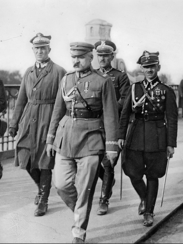 Zdjęcie przedstawia straszego już Piłsudskiego w mundurze, z posiwiałym wąsem. O krok za Marszałkiem idzie kilku oficerów. 