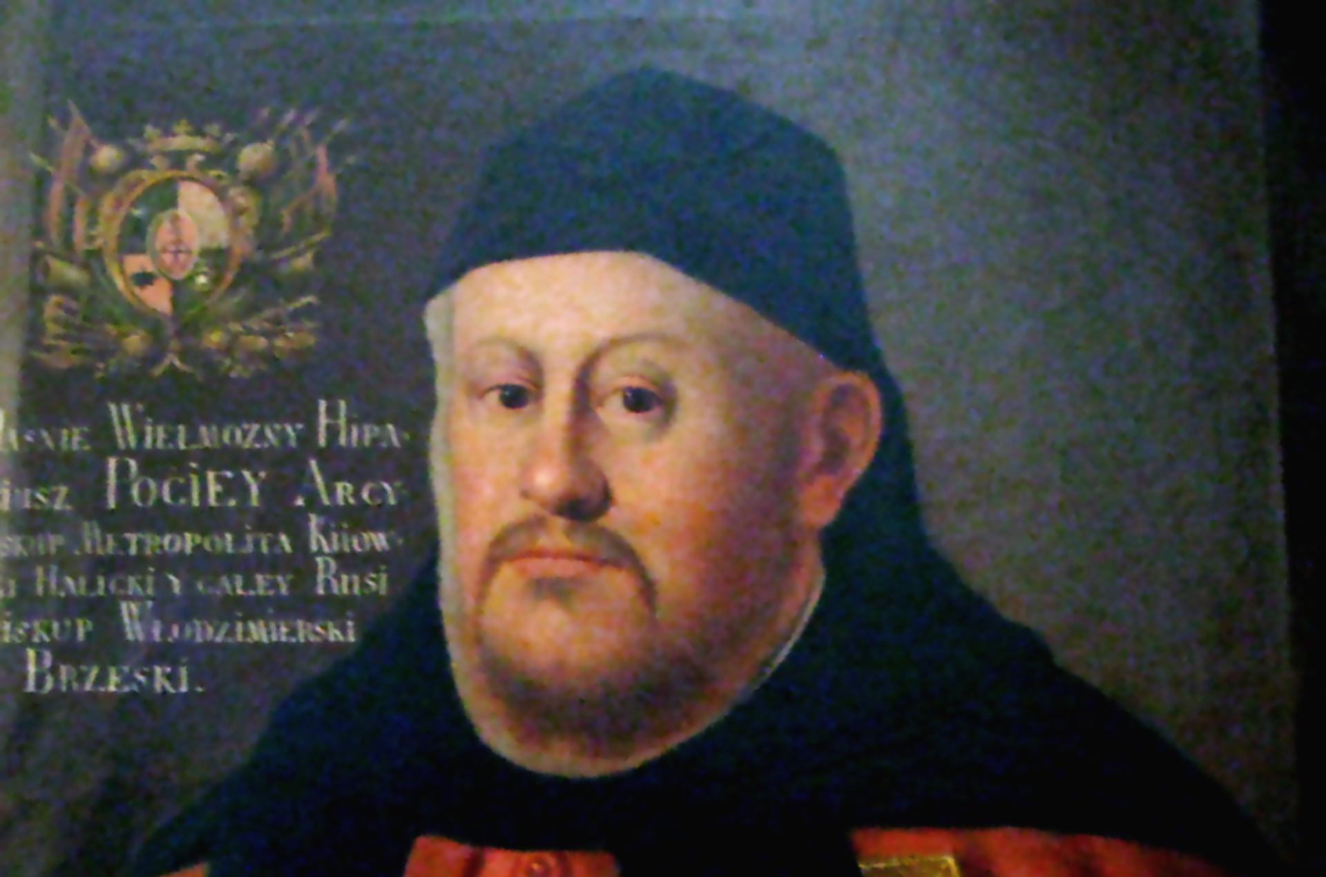 Duchowny w czerwonych szatach i czarnym nakryciu głowy.