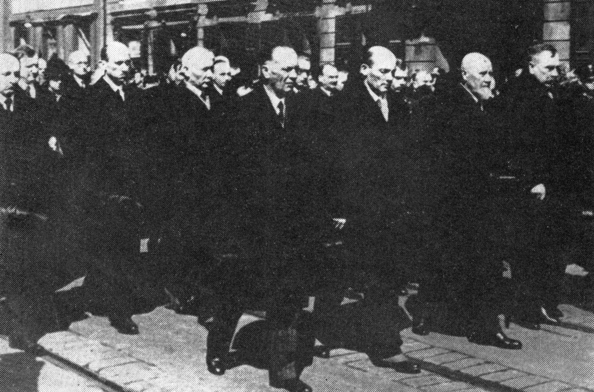 Kondukt żałobny idący ulicą miasta, złożony z mężczyzn ubranych w ciemne garnitury i płaszcze.