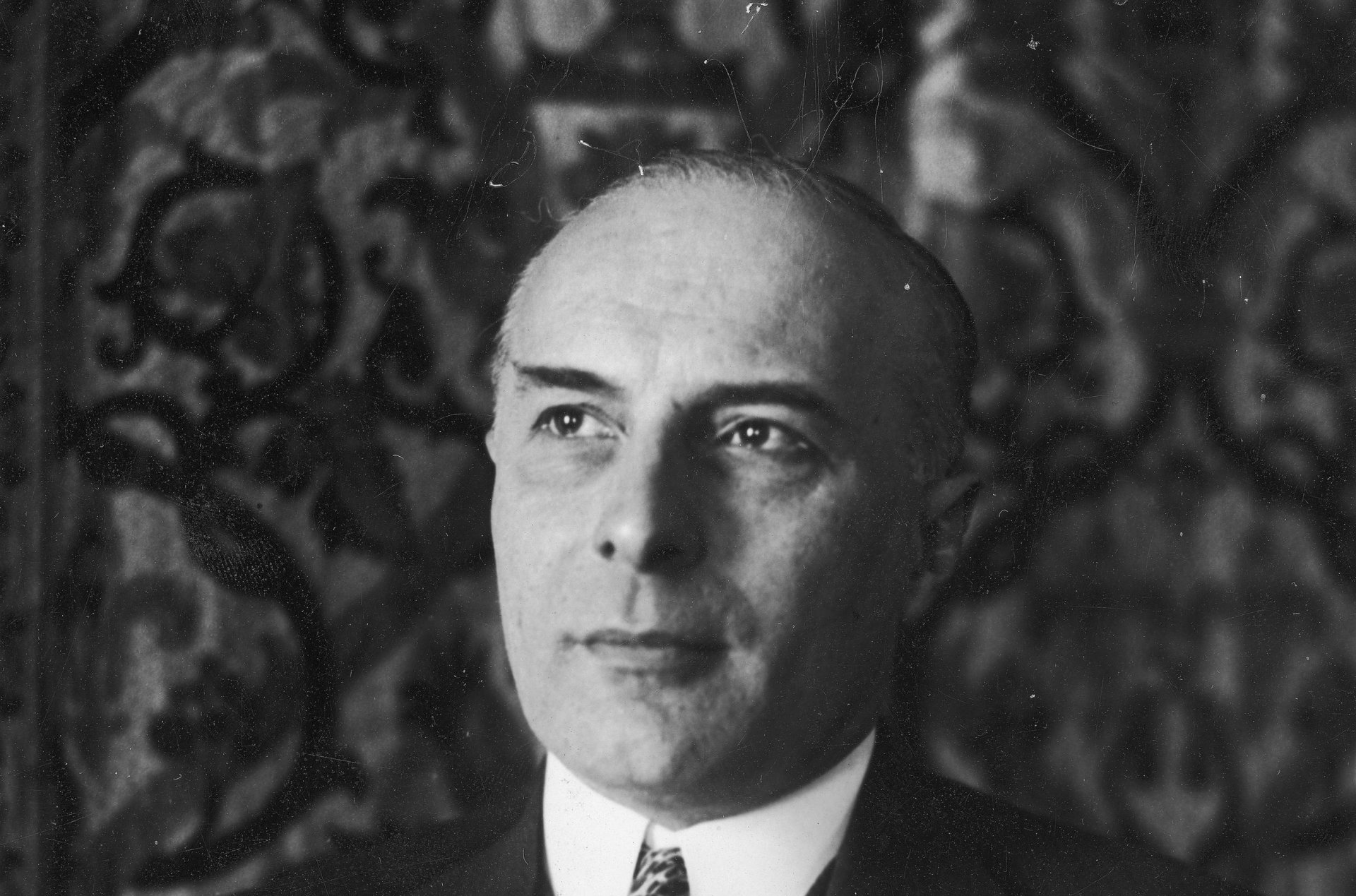 Mężczyzna w średnim wieku w ciemnej marynarce i szerokim krawacie w kratę do białej koszuli.