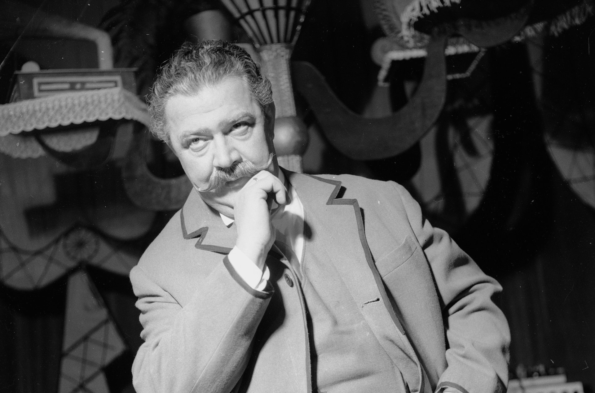 Mężczyzna w jasnym garniturze, z sumiastym jasnym wąsem, stoi na tle dekoracji teatralnych, jedną dłonią podpiera podbródek, kciuk drugiej trzyma w kieszonce kamizelki.