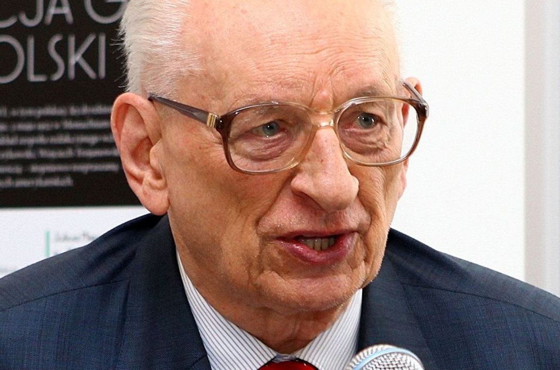 Starszy łysiejący mężczyzna w okularach, granatowej marynarce i czerwonym krawacie. Mówi do mikrofonu.