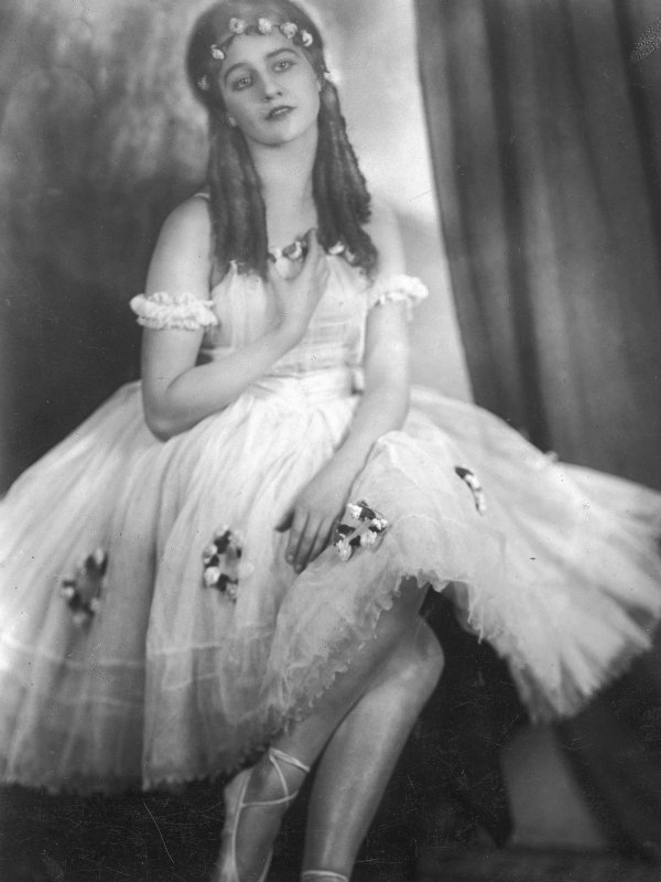 Zdjęcie przedstawia piękną, młodą kobietę w białej sukni tancerki i w baletkach. Kobieta ma zamyśloną twarz, loki sięgające do ramion i wianek na głowie.